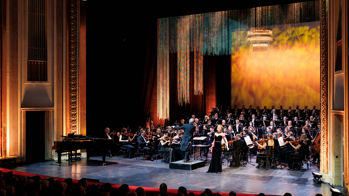 An opera singer performing on stage, with an orchestra behind her. - grafika artykułu