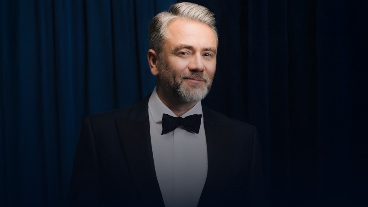 Photo of a smiling Kuba Badach, wearing a tuxedo and bow tie. Dark background.