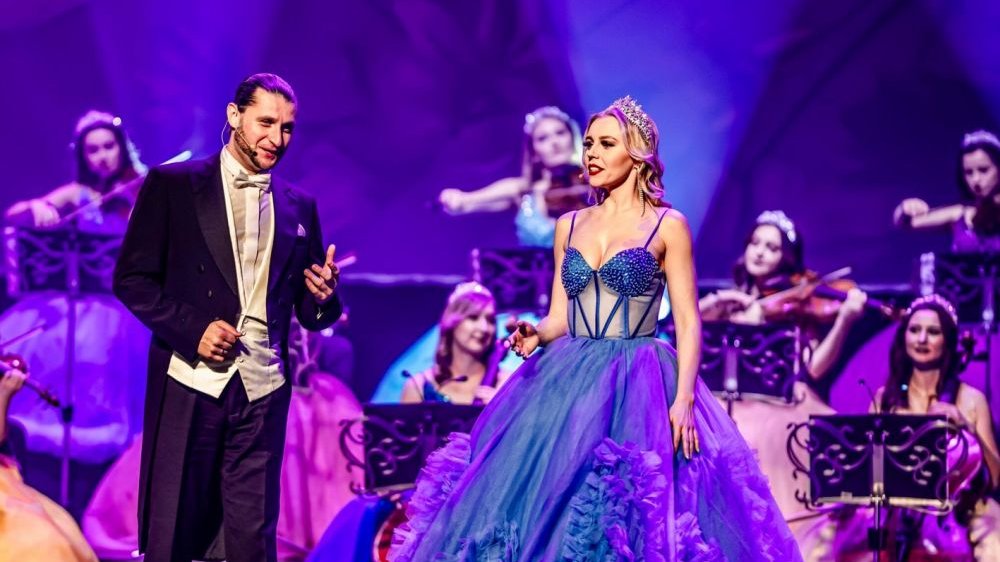A man wearing a tailcoat and a woman in a long ball gown, behind them several women in ball gowns playing instruments.