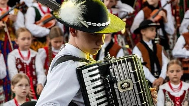 A young boy playing the accordion, in highland costume. In the background, girls and boys in highland folk costumes, some of them hold violins.