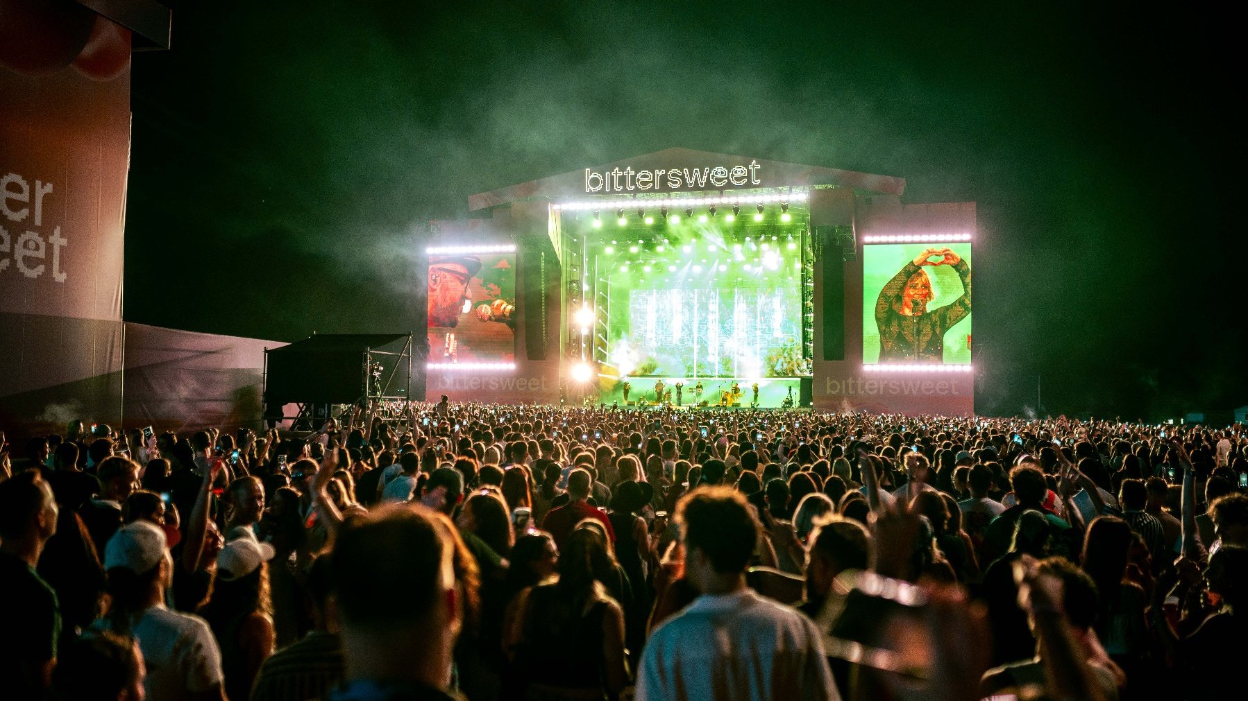 A large, illuminated stage, the band in the spotlight, the audience in front of the stage, and screens on the side of the stage.