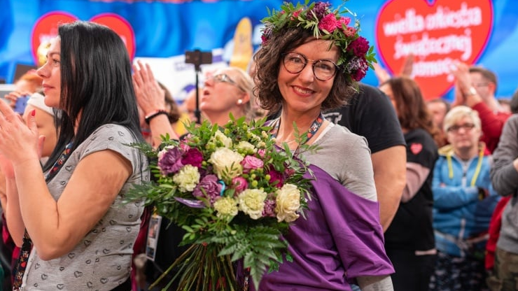 A smiling woman in a flower wreath and with a bouquet of flowers in her hands enjoys the final concert.