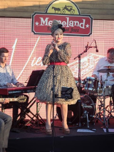 Photo of the performers on stage - a woman in a dress who is standing and holding a microphone in her hands and four men sitting and playing the musical instruments.