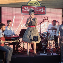 Photo of the performers on stage - a woman in a dress who is standing and holding a microphone in her hands and four men sitting and playing the musical instruments.
