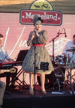 Photo of the performers on stage - a woman in a dress who is standing and holding a microphone in her hands and four men sitting and playing the musical instruments.