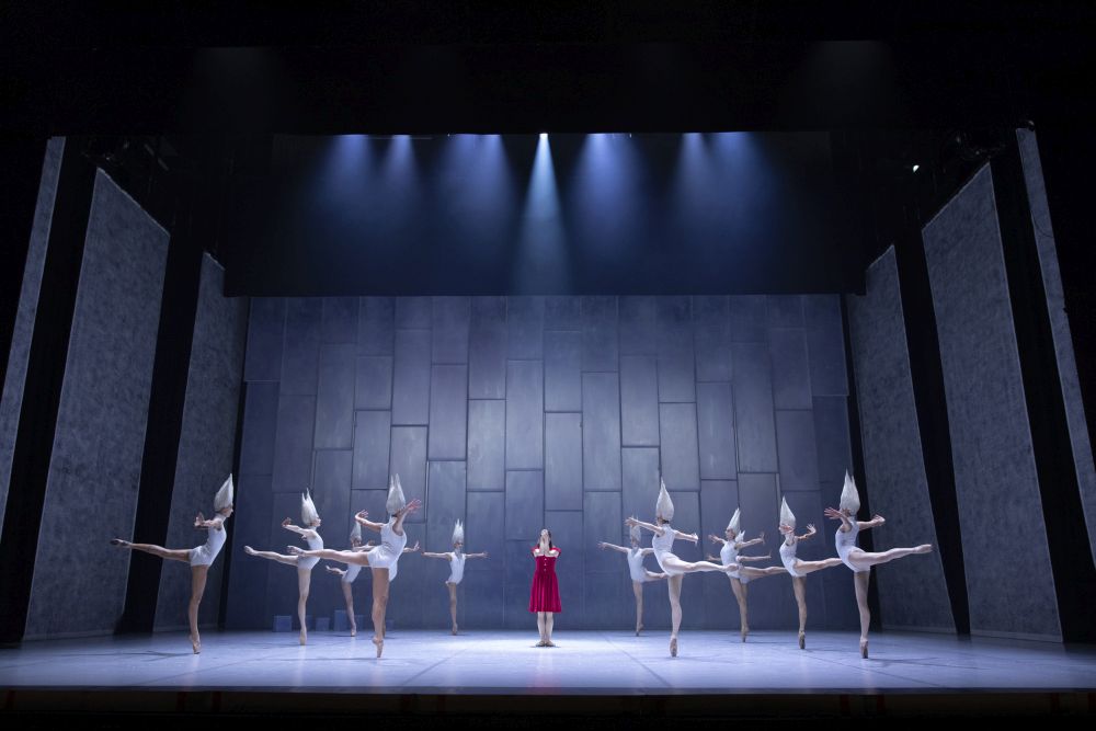 On the stage there are ballerinas in white costumes, and between them in the middle stands a woman in a red dress.