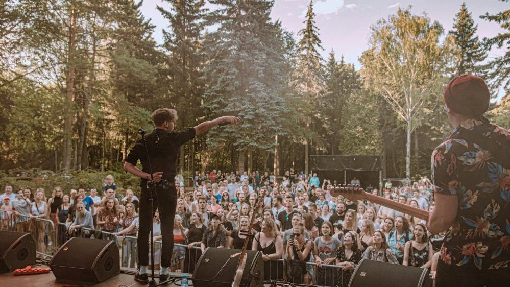 Photo of a man performing on stage. In front of stage people standing and listening to a man. Trees in the background.