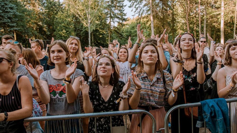 Photo of people standing behind railings, looking up and clapping their hands. Trees in the background.