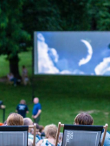 Picture presents some people sitting in a park on deck chairs, watching a film on a big screen