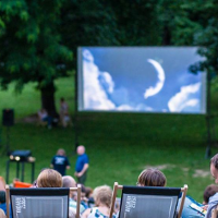 Picture presents some people sitting in a park on deck chairs, watching a film on a big screen