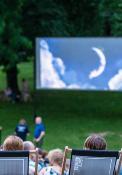 Picture presents some people sitting in a park on deck chairs, watching a film on a big screen