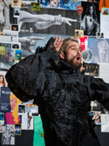 Photo of a man in a black outfit, looking like a fur. In the background various pictures on the wall.