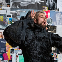Photo of a man in a black outfit, looking like a fur. In the background various pictures on the wall.