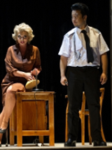 Photo from the performance: a woman sitting on a desk and a man standing next to her.