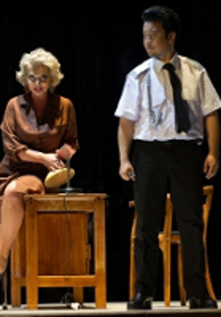 Photo from the performance: a woman sitting on a desk and a man standing next to her.