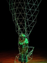 Photo presents an actor in green garment inside a green openwork wire construction on black background