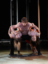 On the stage, a bare-chested man holds two women bent backwards on either side of him. Dark background.