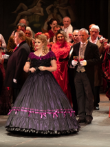 Photo from the spectacle: a woman dressed in a ball gown,many people around her, some of them with wine glasses in their hands.