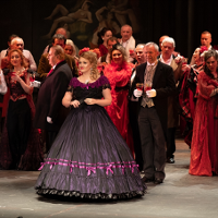 Photo from the spectacle: a woman dressed in a ball gown,many people around her, some of them with wine glasses in their hands.