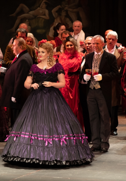 Photo from the spectacle: a woman dressed in a ball gown,many people around her, some of them with wine glasses in their hands.