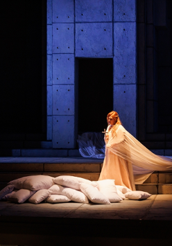 Photo from performance: a woman in a long, white dress standing on stage. She holds a cross in her hand. She stands next to several white pillows on the floor. Black and dark blue background.