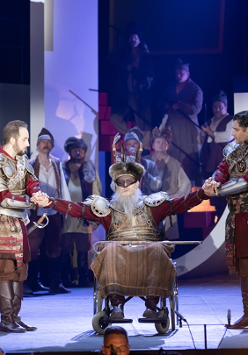 Photo from the performance - three men in armor, one of them sitting between two others, who are standing and holding his hands. People in various costumes in a background.