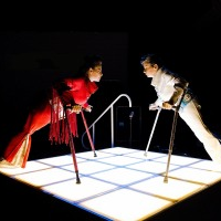 Photo depicts two performers leaning on crutches, standing opposite each other in shiny outfits on square lighted floor.