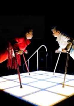 Photo depicts two performers leaning on crutches, standing opposite each other in shiny outfits on square lighted floor.