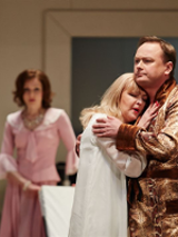 In the foreground there is a couple embracing - a woman in a nightgown, a man in a bathrobe. In the background, a woman in a pink dress looks at them.