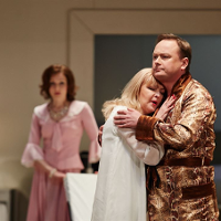 In the foreground there is a couple embracing - a woman in a nightgown, a man in a bathrobe. In the background, a woman in a pink dress looks at them.