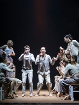 Photo of a group of dancers, some of them standing and some of them sitting on stools.