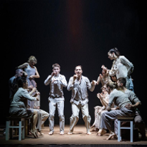 Photo of a group of dancers, some of them standing and some of them sitting on stools.