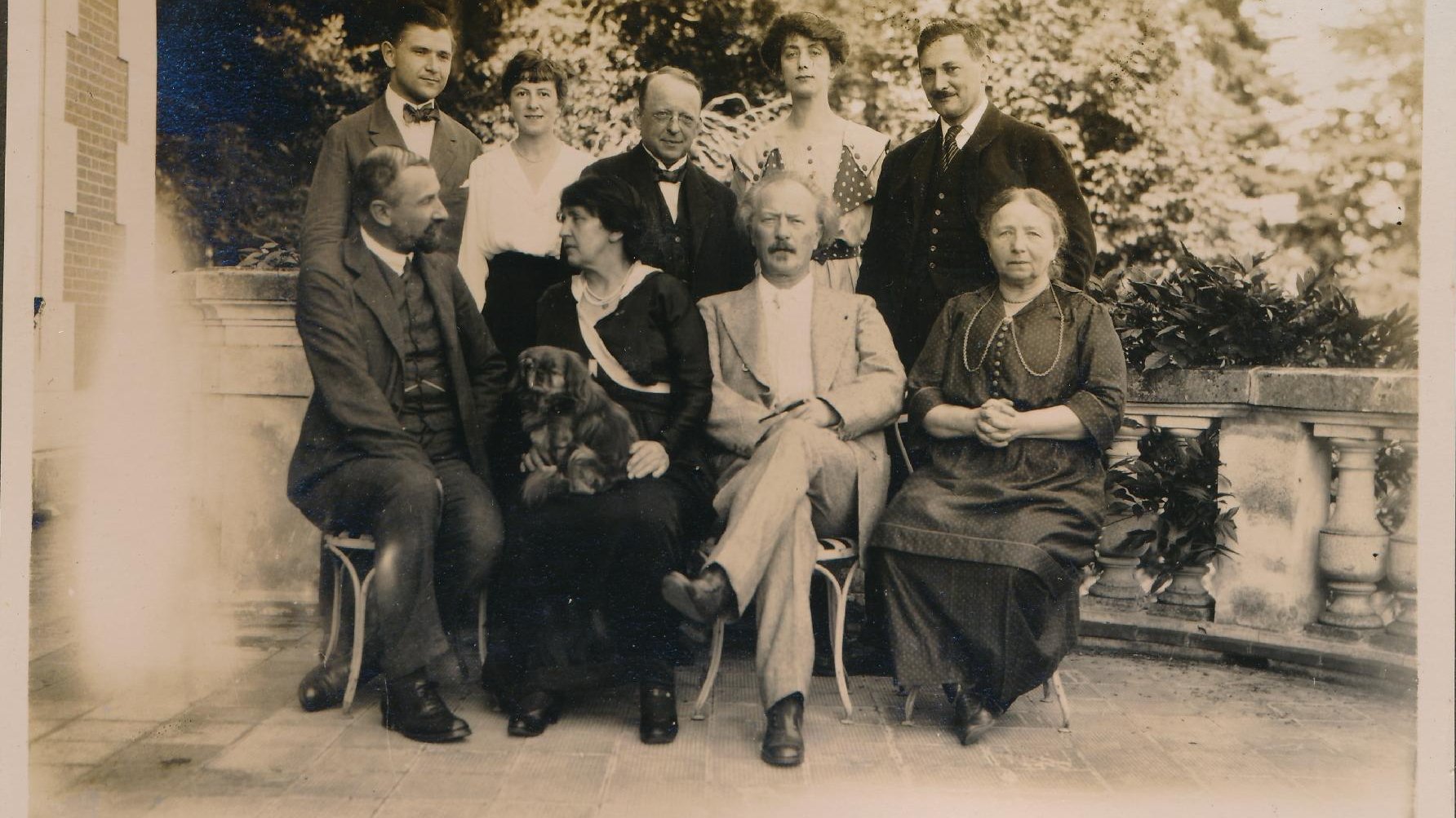 Picture of group of people, posing to the camera. Four of them are sitting, five is standing. The elements of a building and trees in the background.