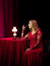 Photo of a woman in red dress, sitting at the table with a glass of champagne. The table is covered by dark red table-cloth, red curtains as a background.