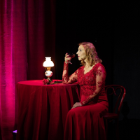 Photo of a woman in red dress, sitting at the table with a glass of champagne. The table is covered by dark red table-cloth, red curtains as a background.