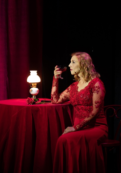 Photo of a woman in red dress, sitting at the table with a glass of champagne. The table is covered by dark red table-cloth, red curtains as a background.