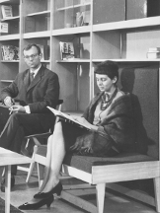 Black and white photo - a woman sitting and reading a book and a man sitting by a table, looking into the camera. In the foreground a small, low table with a ceramic bowl on it. In the background shelves with single books on them.