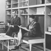Black and white photo - a woman sitting and reading a book and a man sitting by a table, looking into the camera. In the foreground a small, low table with a ceramic bowl on it. In the background shelves with single books on them.