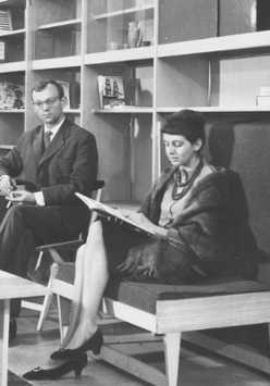 Black and white photo - a woman sitting and reading a book and a man sitting by a table, looking into the camera. In the foreground a small, low table with a ceramic bowl on it. In the background shelves with single books on them.