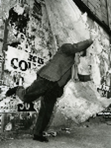 Black and white photo - a man standing on one leg with one arm stretched. A wall with various captions in a background.