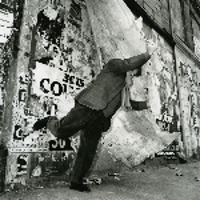 Black and white photo - a man standing on one leg with one arm stretched. A wall with various captions in a background.
