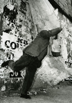 Black and white photo - a man standing on one leg with one arm stretched. A wall with various captions in a background.