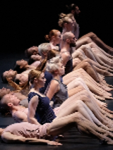 Photo of dancers in a row - some of them lying on the floor, some of them sitting on the floor. Black background.