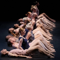 Photo of dancers in a row - some of them lying on the floor, some of them sitting on the floor. Black background.