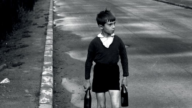 Black and white picture of a boy walking along a road and holding two bottles in his hands. He is dressed in old-fashioned clothes.