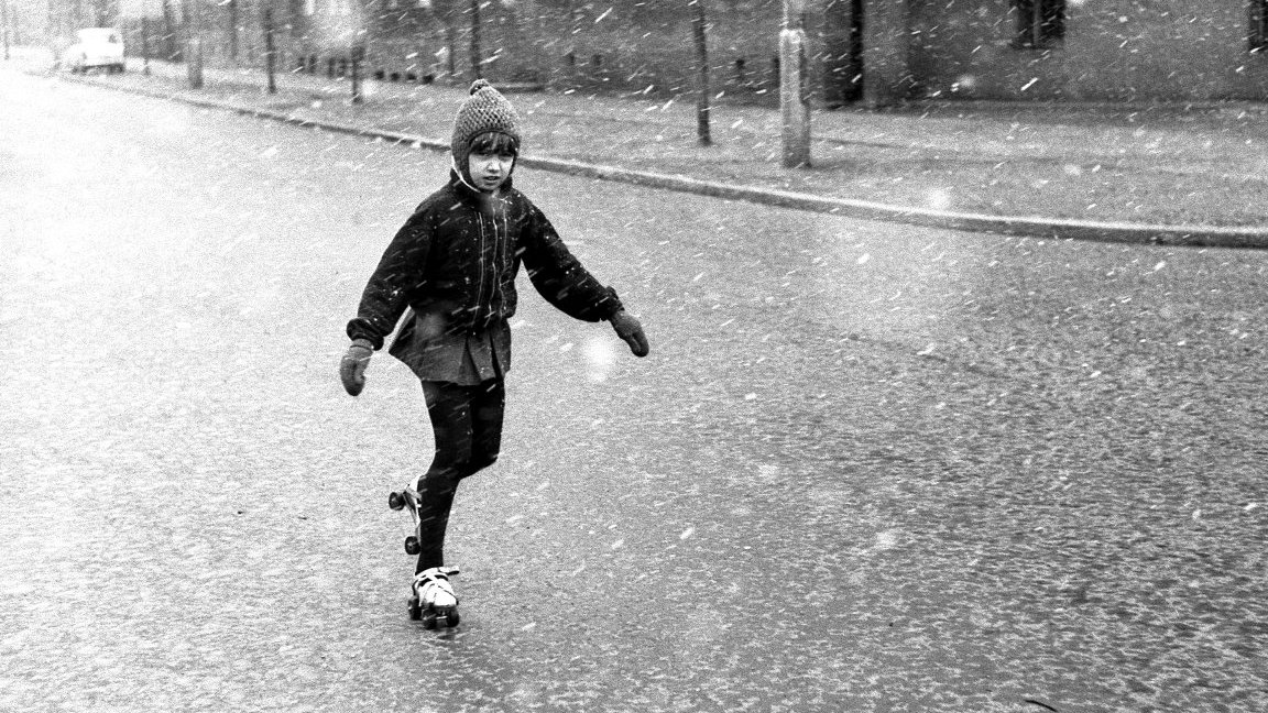 Black and white photo of a roller-skating girl on a street. The girl is wearng warm clothes, it is snowing.