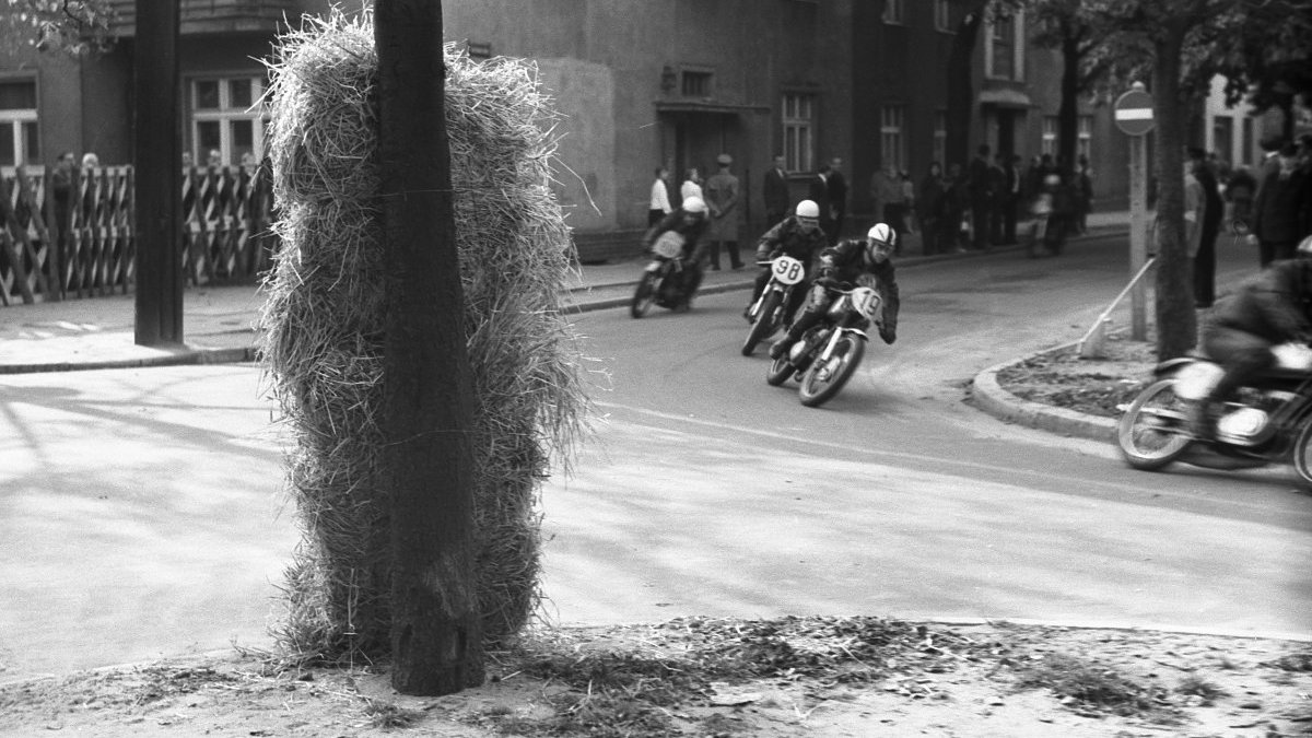 Black and white picture of motorcyclists taking part in a street ride. In the background some observers of a ride.