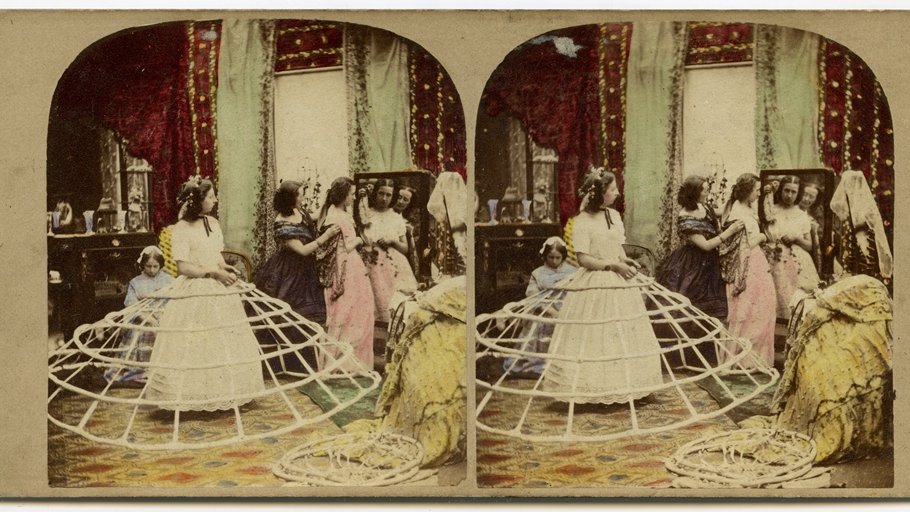 Stereoscopic photograph presenting three women: two of them are looking at the mirror, one of them is standing in the middle of a room wearing frame of a crinoline
