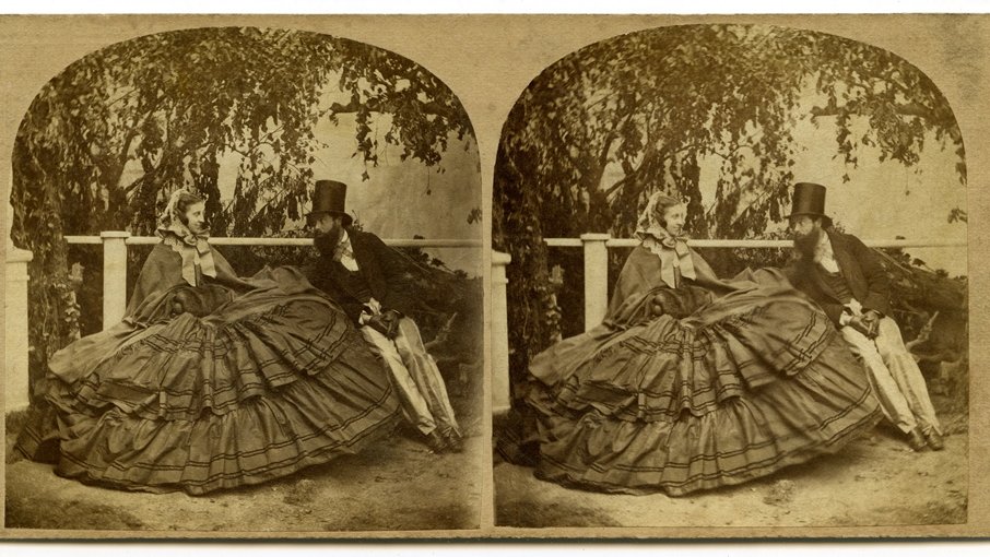 Stereoscopic photograph in sepia colours - a man and a woman in a crinoline sitting on a bench. A man is leaning towards a woman, they are looking at each other. In the background some trees and bushes.