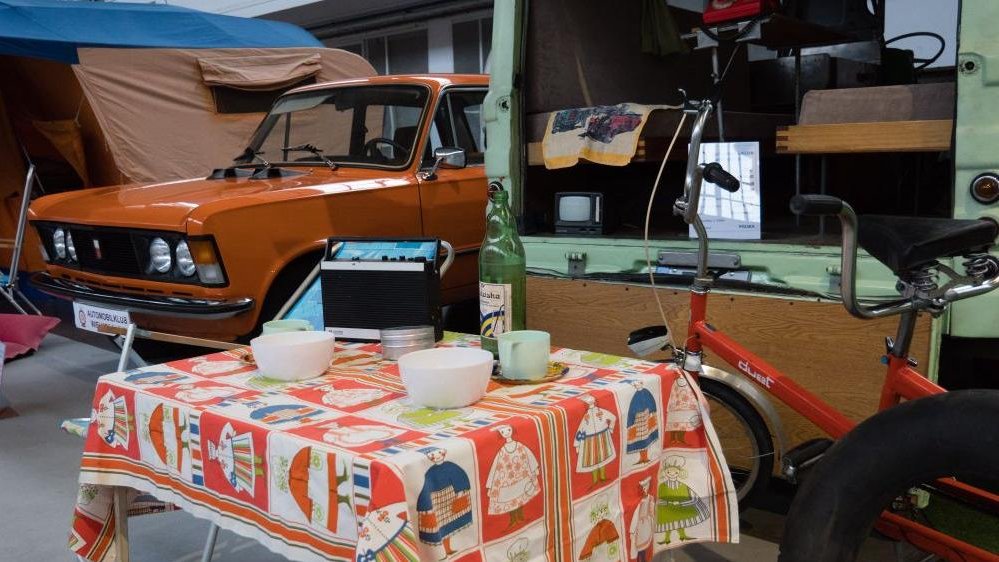 The photo from the exhibition: the exhibits from PRL period - a table with oilcloth, a bottle, a radio and a few dishes on it, a bicycle by te table and a car from PRL period in the background.